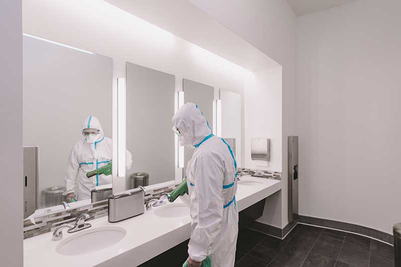 Professional commercial cleaner cleaning office bathroom near sink area in front of a mirror in full body covered suit