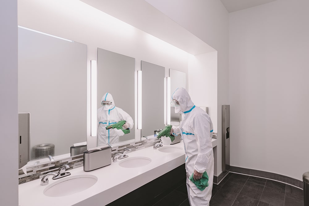 Professioanl commercial cleaner in all white suit cleaning sinnk area of corporate office in front of a mirror