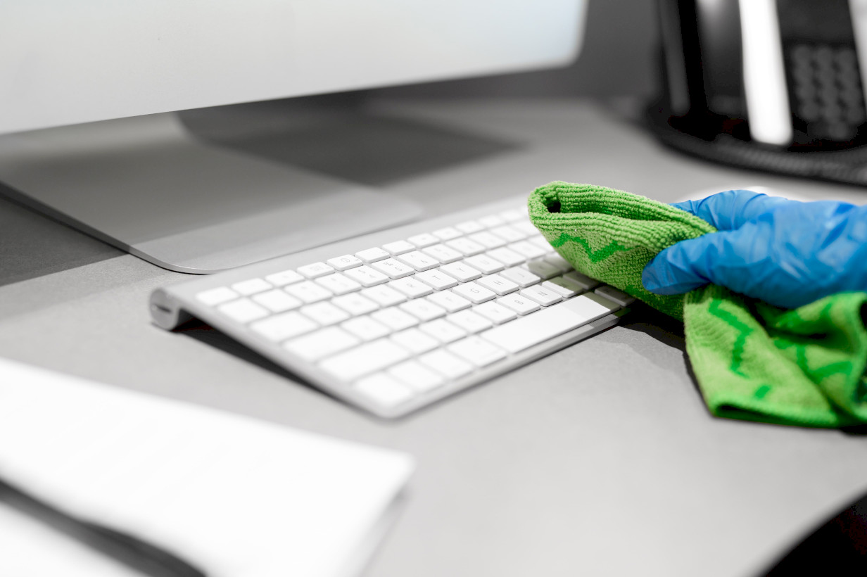 professional cleaners hand in blue glove using green rag to wipe desk space around computer in effort to reduce risk of spreading flu